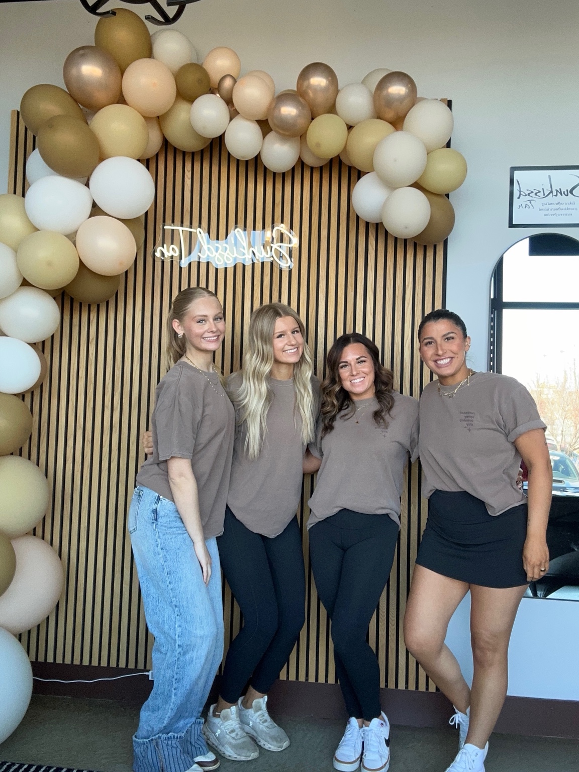 Four Sunkissd Tan team members smiling in front of the salon’s wood feature wall